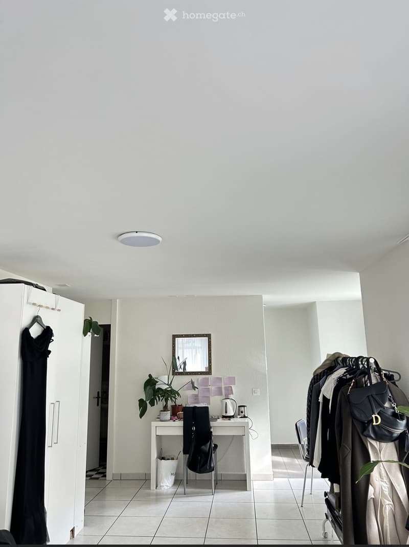 A modern, minimalist room with a white tiled floor, white walls, and a large ceiling light. Features include a small desk with a chair, a mirror, a potted plant, a coat rack with several garments, and a white cabinet with a hanging dress.
