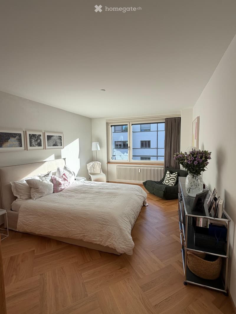 Bright bedroom with wooden flooring, large window with curtains, bed with white bedding, side table with lamp, armchair, and decorative vase on a small table.