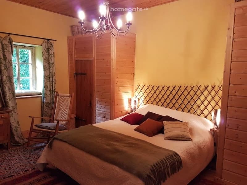 Cozy bedroom with wooden walls, a large bed with pillows and a blanket, a rocking chair, a nightstand with a lamp, and a wooden wardrobe.
