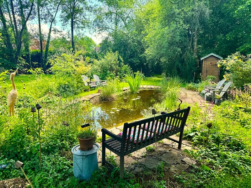 Lush garden with a pond, wooden bench, potted plants, decorative bird sculpture, and outdoor seating area surrounded by greenery and trees.