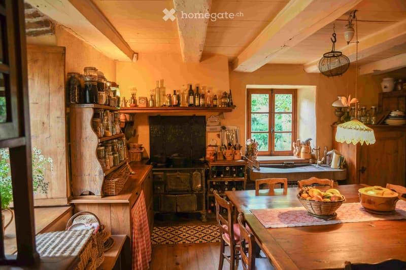 Cozy kitchen with wooden beams, exposed wooden ceiling, and rustic decor. Features a wooden dining table with chairs, a stove, shelves with jars and bottles, and a sink with a window above.