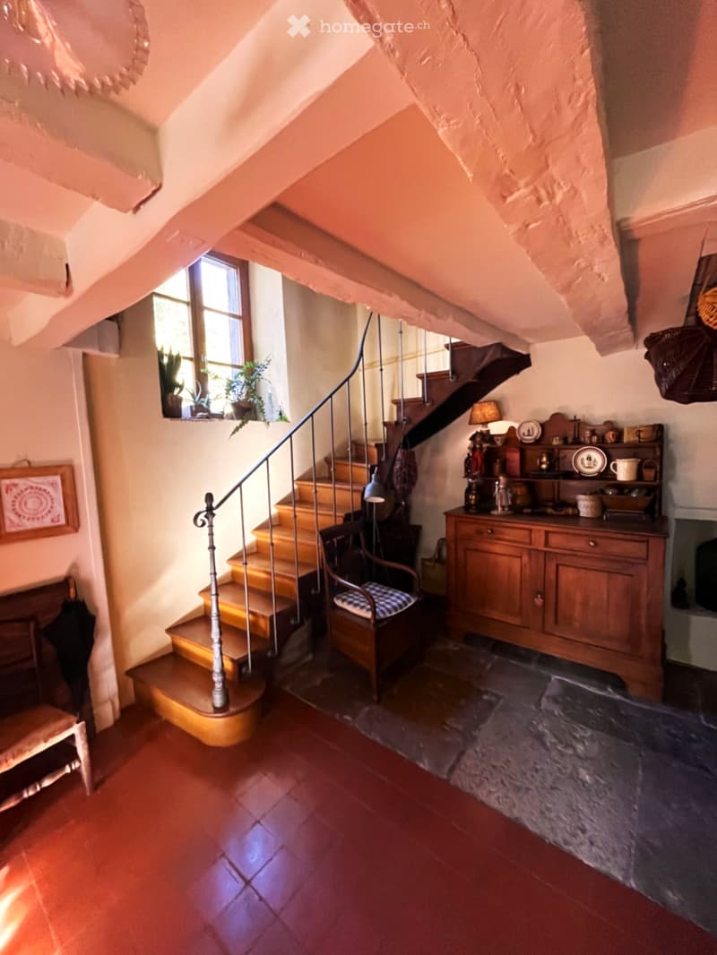 Staircase with wooden steps and a metal railing, leading to an upper level. The hallway features exposed wooden beams, a window with potted plants, a wooden cabinet with decorative items, and a small bench with a cushioned seat.