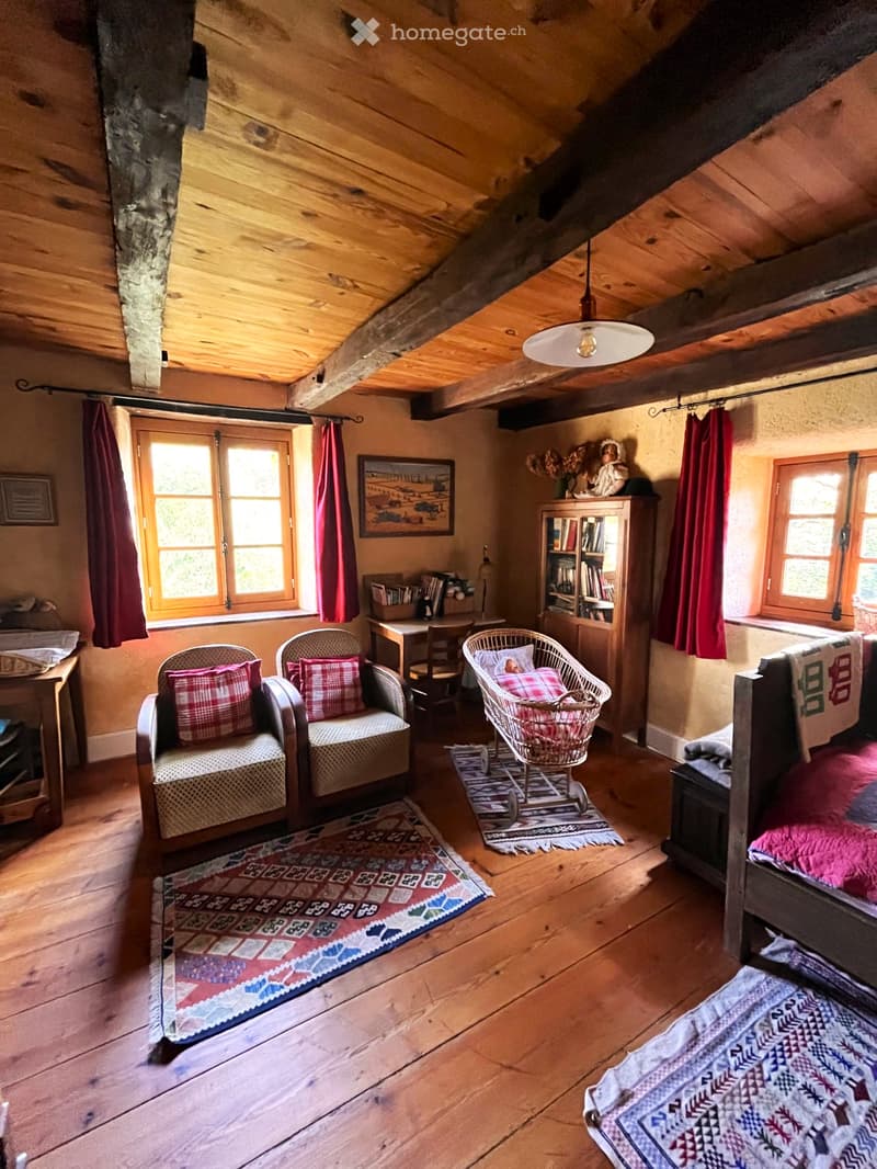 Cozy attic room with wooden beams, exposed stone wall, wooden floor, large windows with red curtains, sofa, armchair, baby chair, bed, bookshelf, and artwork.