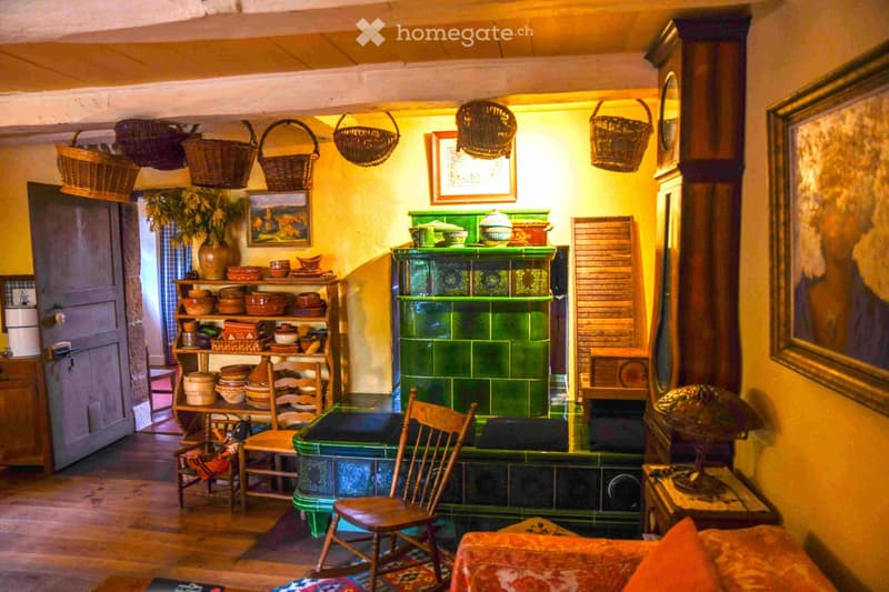 A cozy, rustic room with warm lighting, wooden furniture, and traditional decor including a green tiled stove, woven baskets, and shelves filled with ceramic dishes.