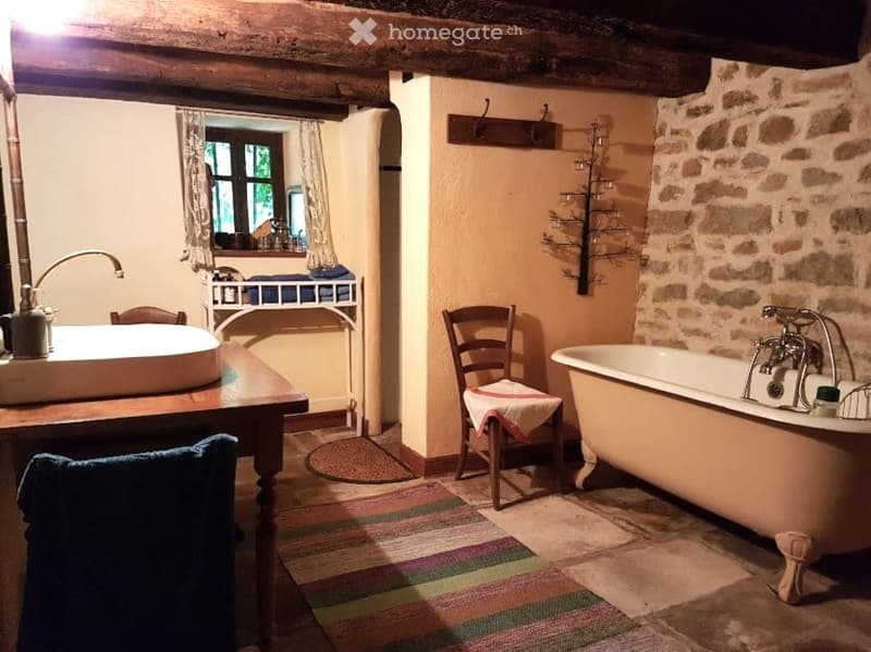 Rustic bathroom with exposed wooden beams, stone walls, a clawfoot bathtub, a wooden sink, a small table, and a chair with a towel draped over it. A window with curtains provides natural light.