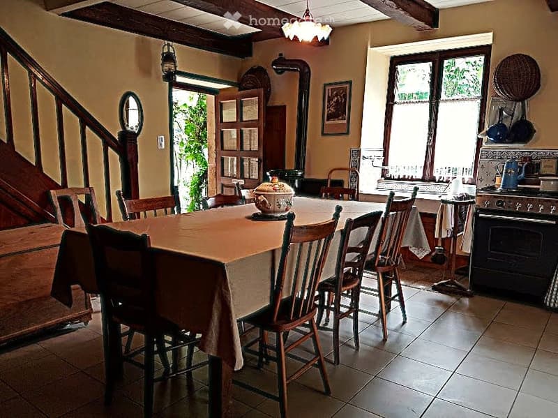 A cozy kitchen with a dining area, featuring a wooden dining table with chairs, a stove, and a framed picture on the wall. Natural light streams in from a large window, and a wicker basket hangs on the wall.