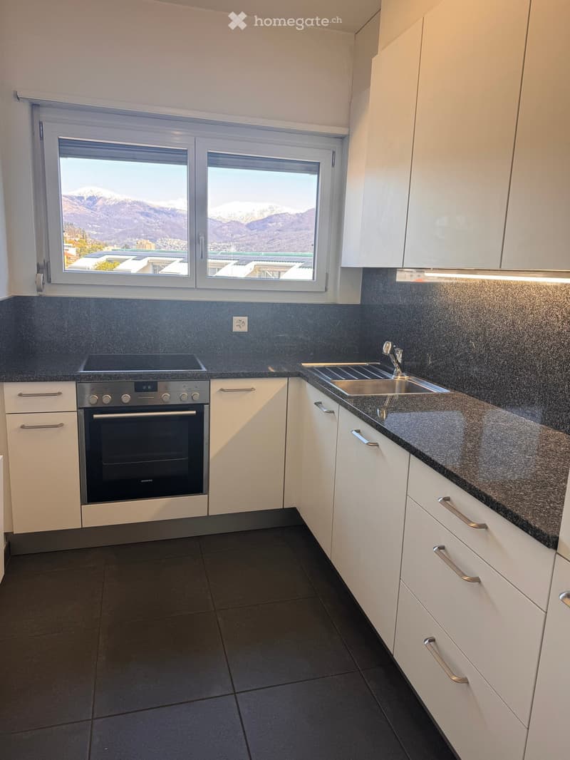 Modern kitchen with white cabinets, dark countertops, stainless steel oven, and a sink with a faucet. Large windows offer a scenic mountain view.