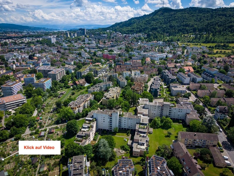 Aerial view of a cityscape with numerous residential and commercial buildings, surrounded by green spaces and trees, with distant mountains visible in the background.