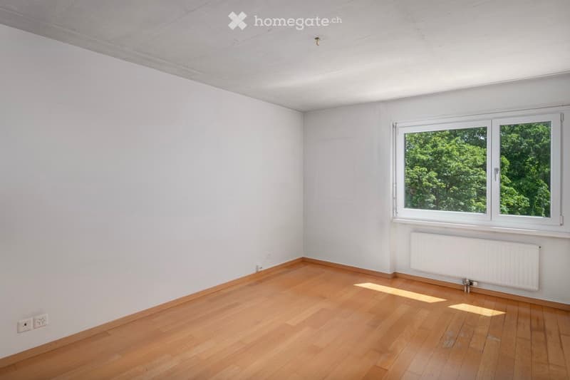 Empty room with light wooden flooring, white walls, and a large window showing greenery outside. No furniture or equipment present.