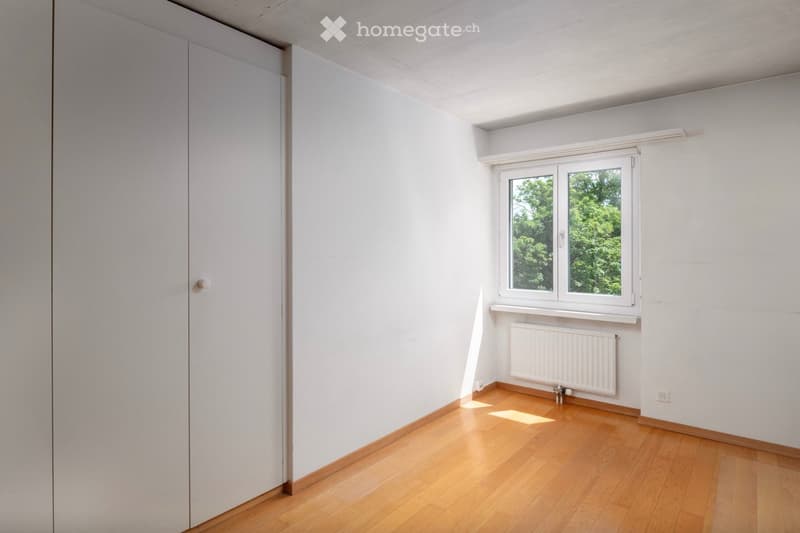 Empty room with light-colored wooden flooring, white walls, a window with white frame, and a radiator below the window.