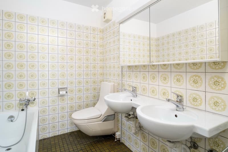 Bathroom with white tiled walls featuring floral patterns, a bathtub, toilet, two sinks, and a mirror above the sinks.
