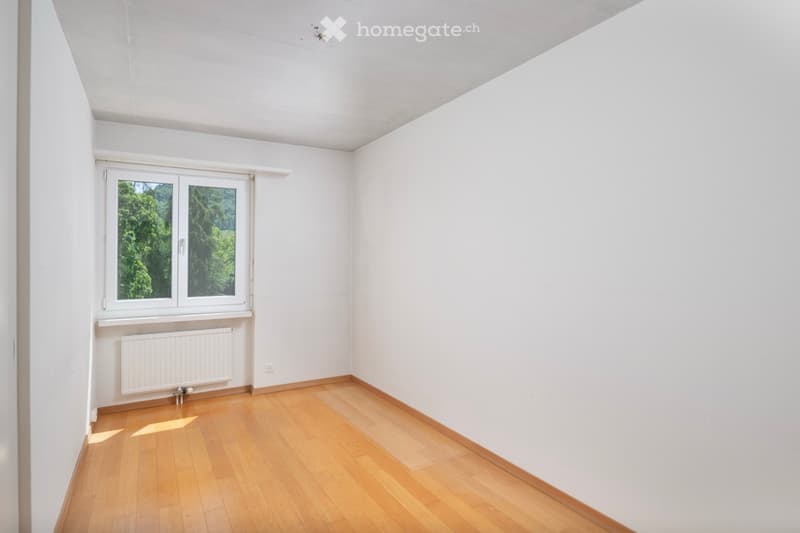 Empty room with white walls, wooden floor, and a window with a view of greenery.