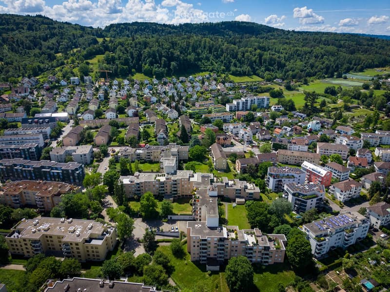 Aerial view of a residential area with multiple apartment buildings, houses, and green spaces surrounded by forested hills.