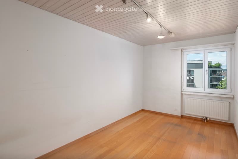 Empty room with wooden flooring, white walls, and a large window with a view of a building and greenery outside.