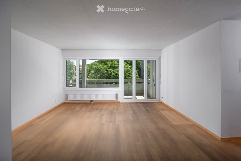 Empty room with wooden flooring, white walls, large windows, and a sliding glass door leading to a balcony.