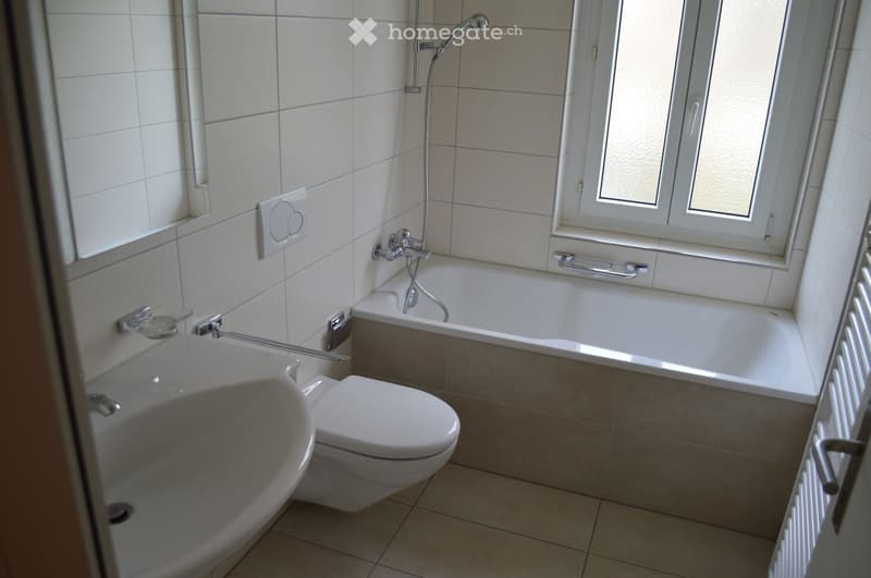 Modern bathroom with a bathtub, toilet, sink, and showerhead. The room features tiled walls and a window with frosted glass.