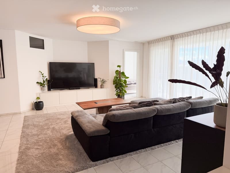 Modern living room with a grey sectional sofa, wooden coffee table, large flat-screen TV, and several potted plants. The space features large windows with sheer curtains and a round ceiling light.
