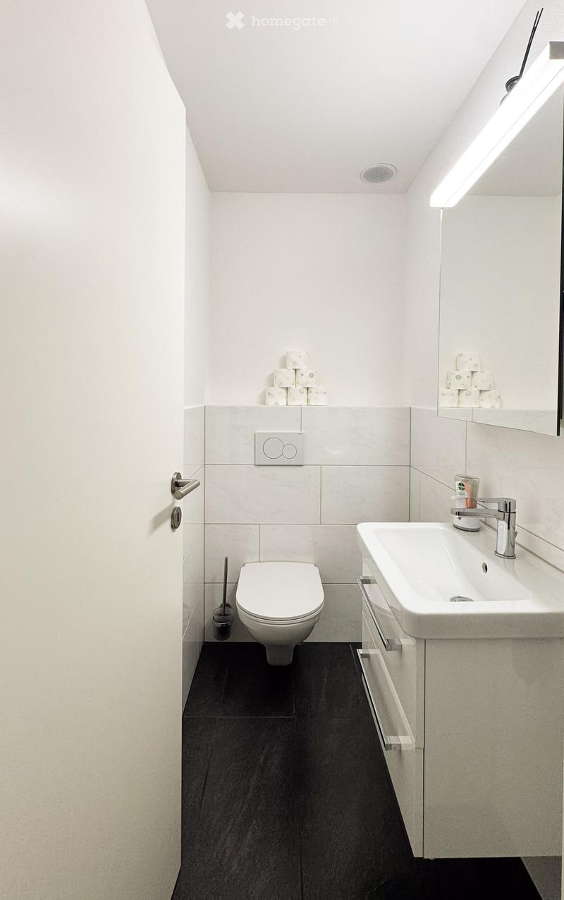 Modern bathroom with white tiles, black floor, toilet, sink, mirror, and toilet paper on shelves.