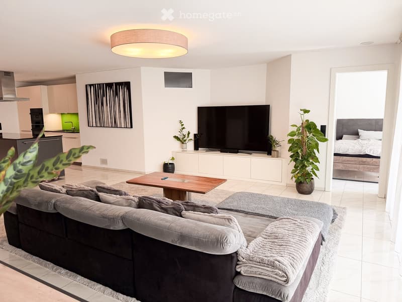 Modern living room with a grey sectional sofa, wooden coffee table, large flat-screen TV, kitchen area with cabinets and island, and a visible bedroom with a bed.