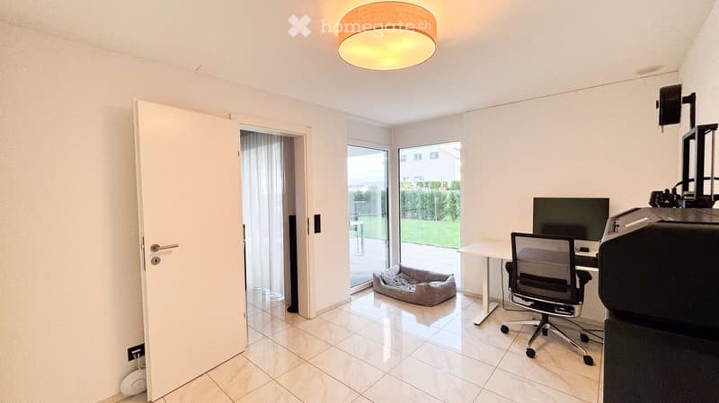 Bright room with tiled floor, white walls, a desk with a chair, a screen, a printer, and a dog bed near the sliding glass door leading to a patio.