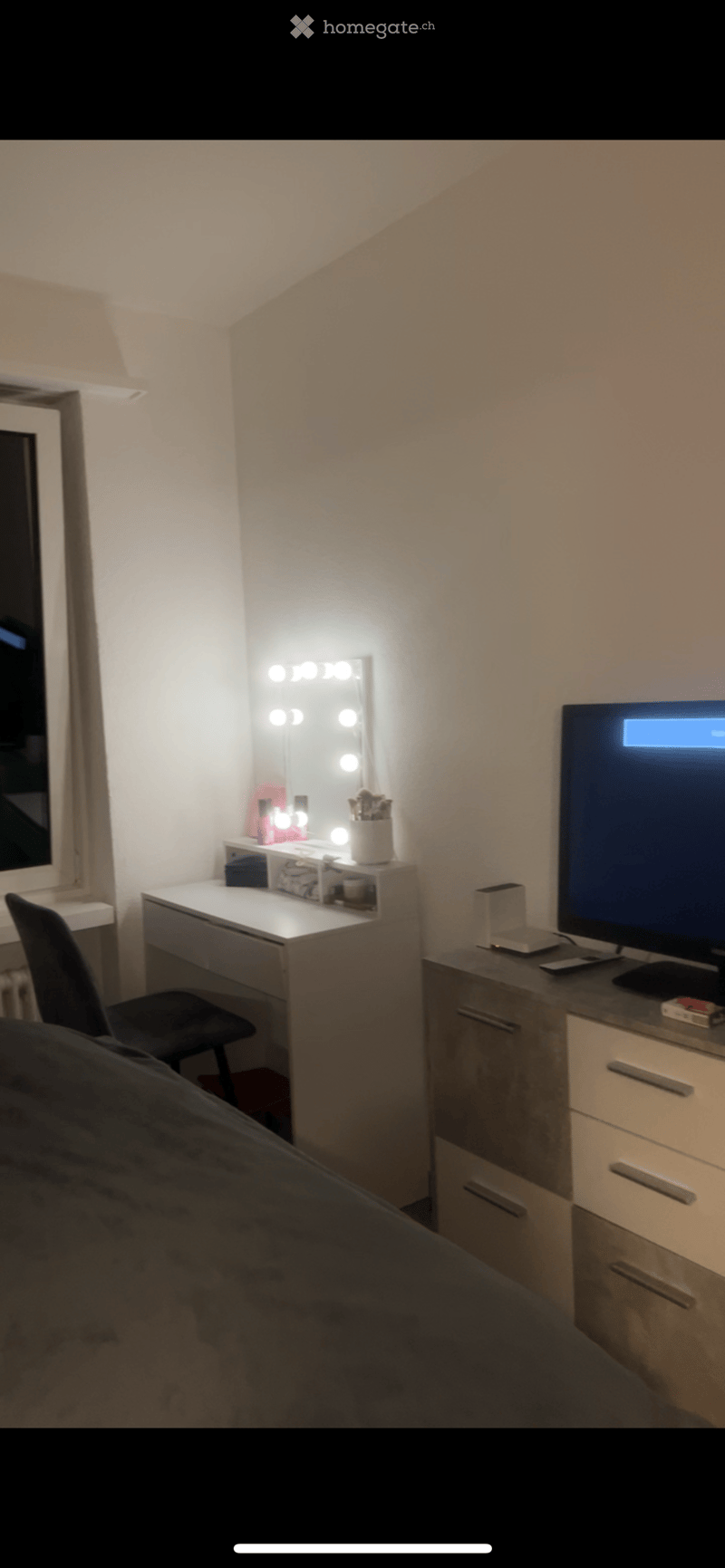 A cozy bedroom with a large mirror, vanity desk, chair, television, and drawer unit. The room features soft lighting and a comfortable bed.