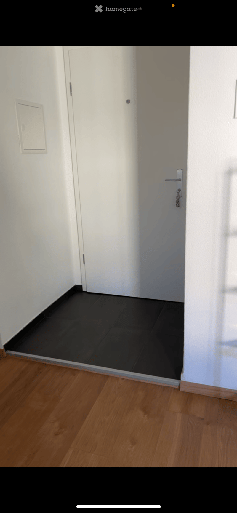 Empty hallway with wooden flooring and white walls, featuring a closed white door and a partially visible adjacent room with black tiled flooring.