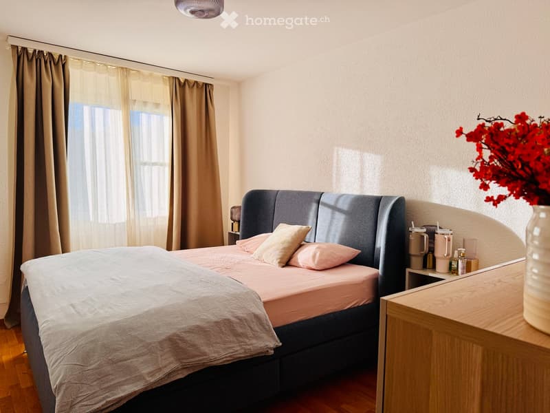 A modern bedroom with a large bed, grey headboard, pink bedding, nightstand, and decorative vase with red flowers on a wooden side table.