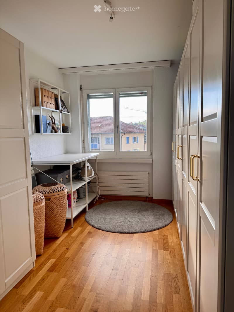Bright hallway with wooden flooring, white walls, and a round grey rug. Features a white shelving unit with storage boxes, a small table, and two woven baskets. A large window at the end offers a view of the street.
