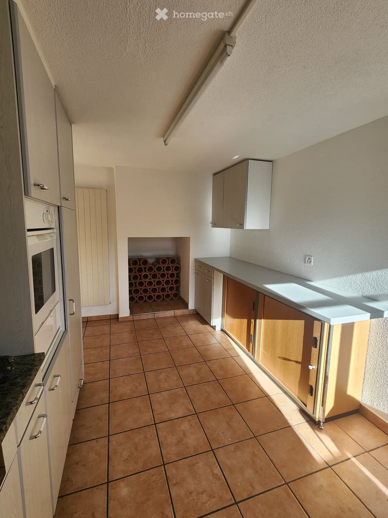 Modern kitchen with tiled floor, light wooden cabinets, and a built-in oven. A wine rack is visible in the background.