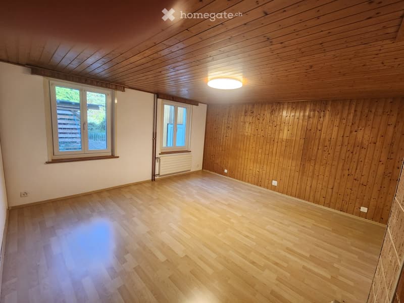 Empty room with wooden flooring, wooden panel walls, and large windows allowing natural light. The ceiling is also wooden with a central light fixture.