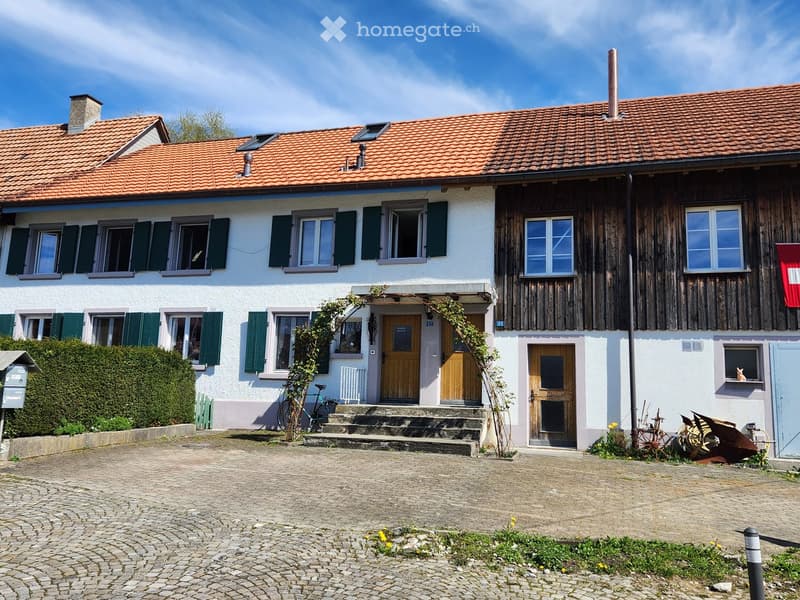 Two-story house with red-tiled roof, white and wooden facades, multiple windows with shutters, and a small garden area.