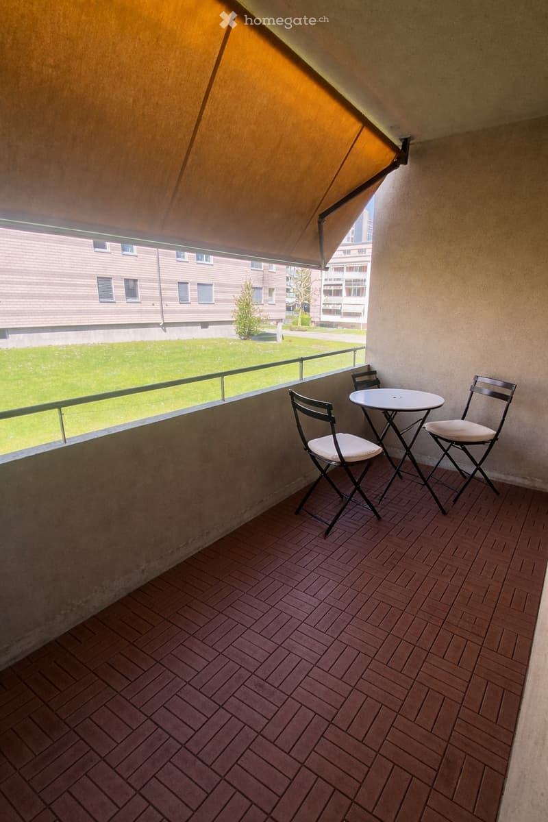 Balcony with wooden decking, small round table, and two folding chairs, overlooking a green lawn and neighboring buildings.