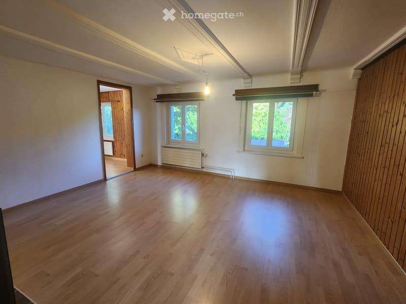 Empty room with wooden flooring, white walls, and large windows with blinds. A door leads to another room, and a radiator is visible under one window.