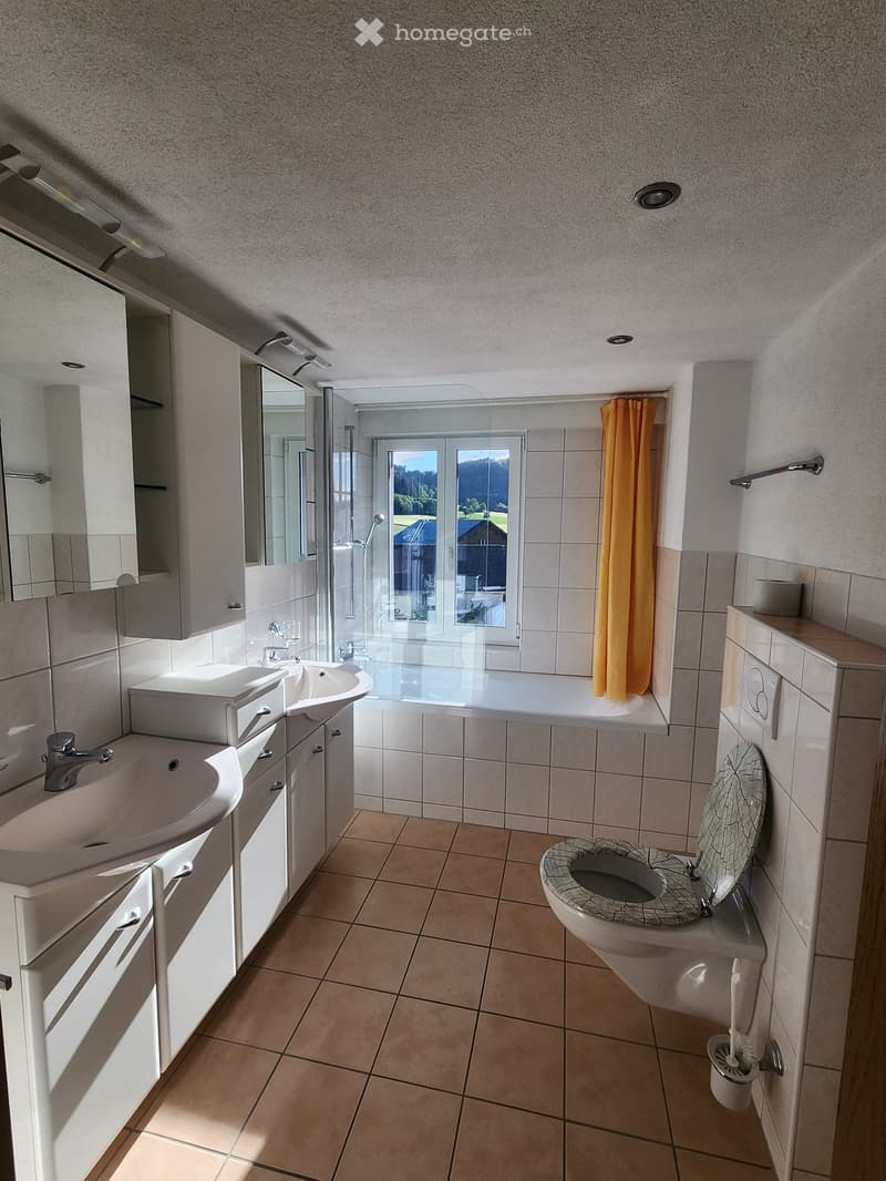 Modern bathroom with double sink, mirror, bathtub, toilet, and a window with a view of greenery.