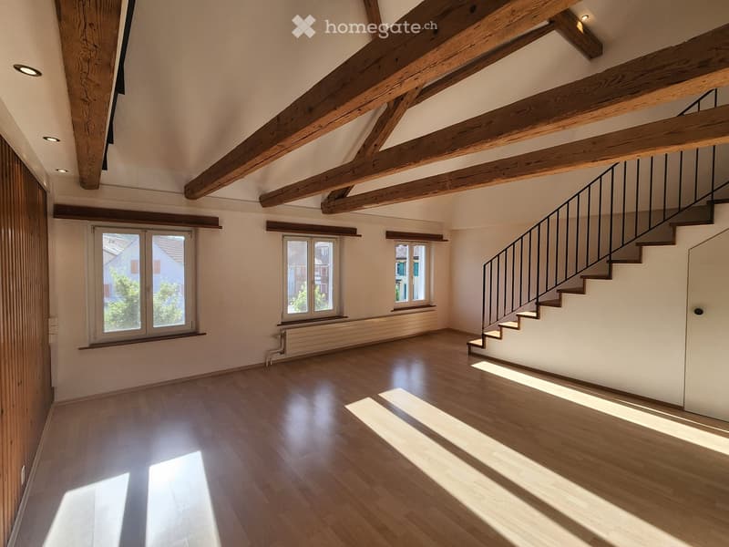 Empty room with wooden floor, exposed wooden beams, large windows, and a staircase leading upwards.