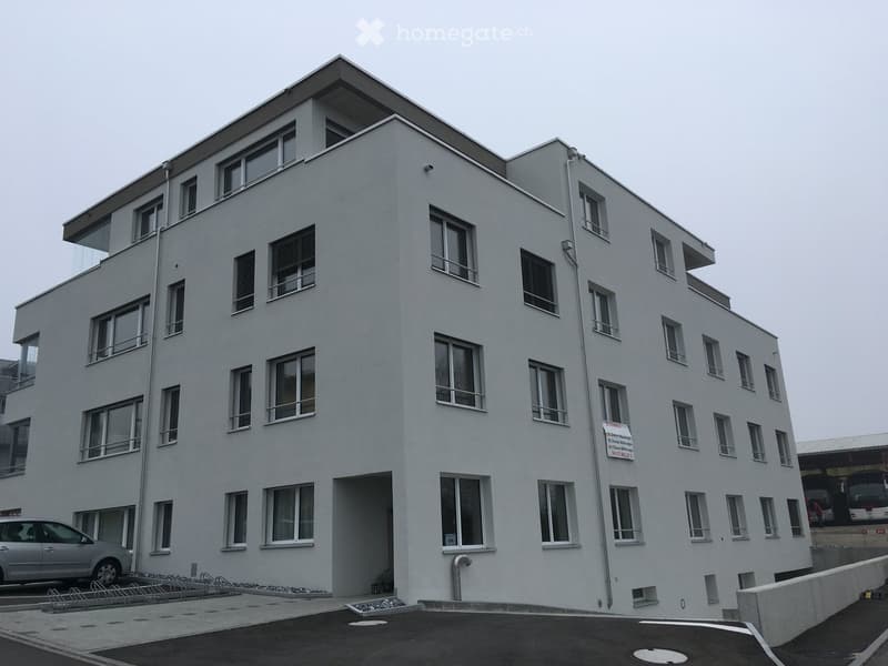 Modern multi-story building with white facade, numerous windows, and balconies. Features a parking area with a car and bicycles.