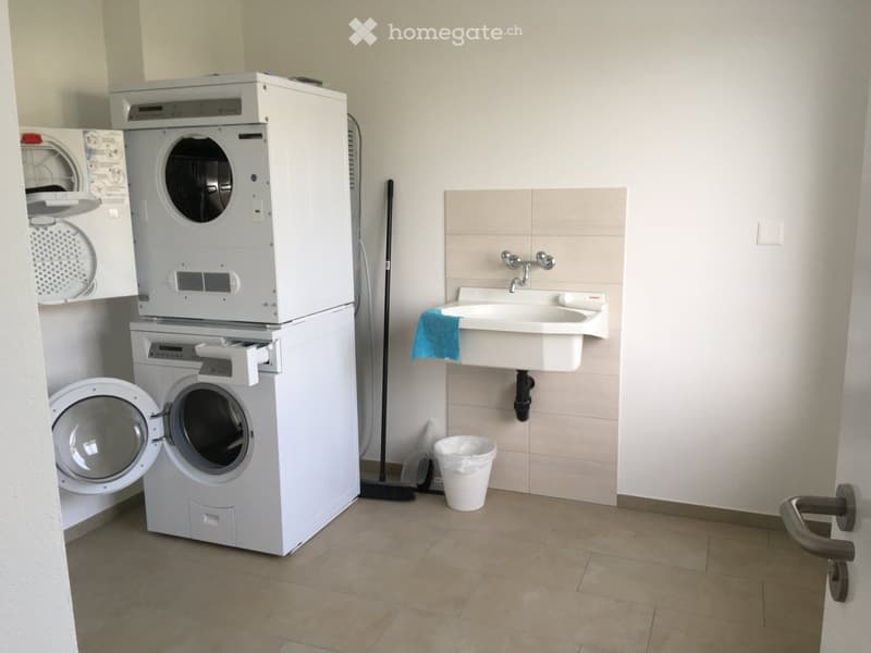 Bathroom with a washing machine and dryer stacked, a sink with a faucet, a towel, a trash can, and a broom.