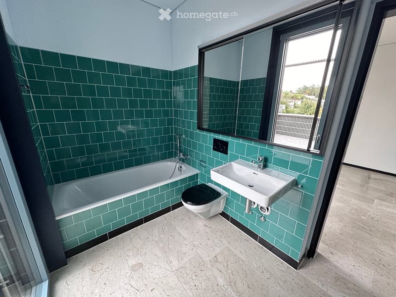 Modern bathroom with green tiled walls, a bathtub, a sink, a toilet, and a large mirror. The room has a large window offering a view of the outdoor area.