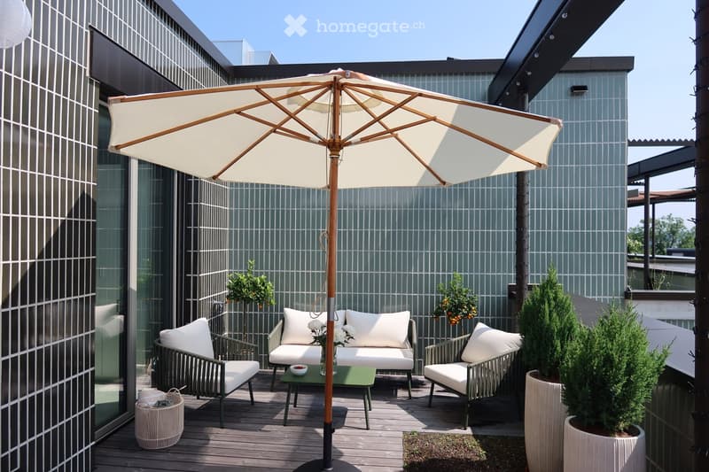 A modern rooftop terrace with a wooden deck, featuring a large umbrella, a sofa set with white cushions, a small green table, and potted plants.