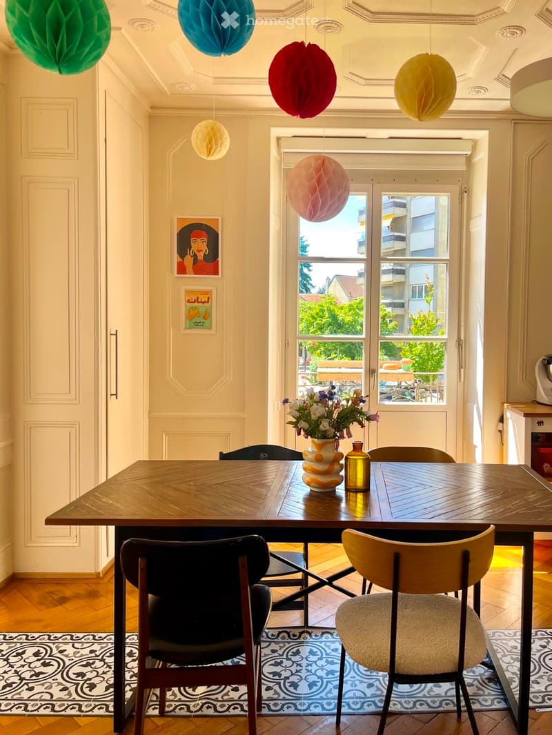 Dining area with wooden table, chairs, vase with flowers, and decorative items. Large windows provide natural light and a view of the street.