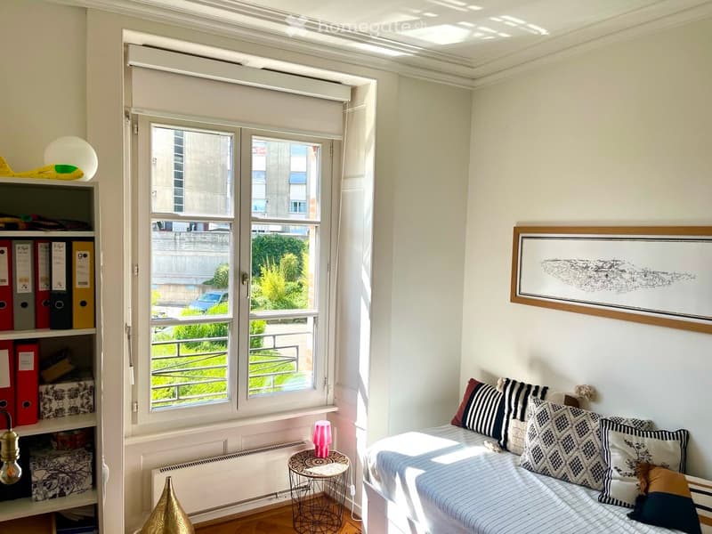 A small, well-lit room with a sofa, a small round table with a pink lamp, a shelf with binders, and a framed picture on the wall. The room has large windows with blinds and a view of greenery outside.