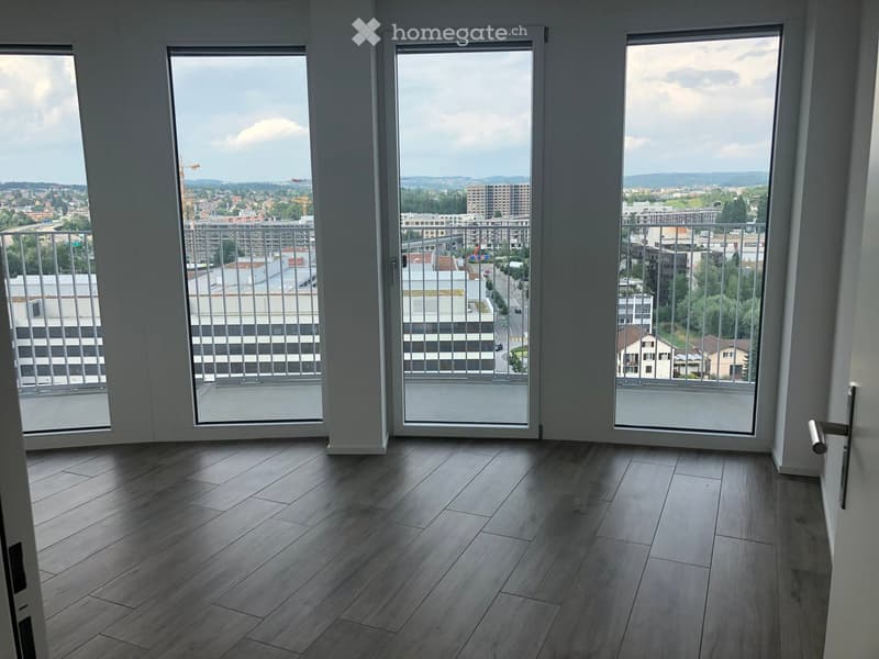 Empty room with wooden flooring and large windows offering a cityscape view. The space is unfurnished, with white walls and modern sliding doors leading to a balcony.