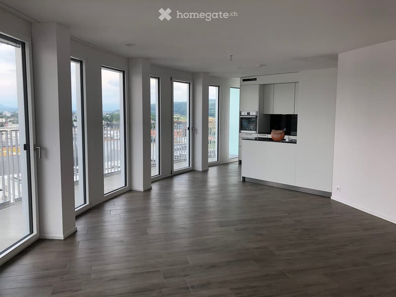 Modern, open-plan living and kitchen area with dark wooden flooring, white cabinetry, and large glass doors leading to a balcony with scenic views.