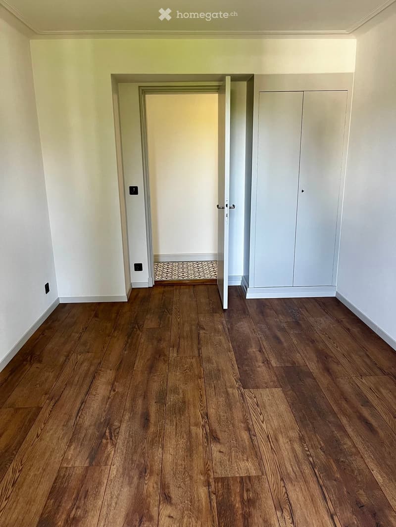 Empty room with wooden flooring, white walls, and a built-in closet with double doors. A small patterned rug is visible in the adjacent room through an open doorway.