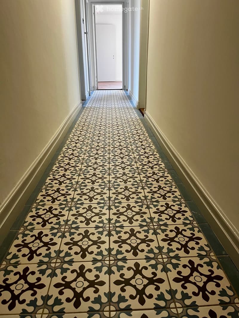 A long hallway with patterned tiles, cream-colored walls, and a visible door at the end.
