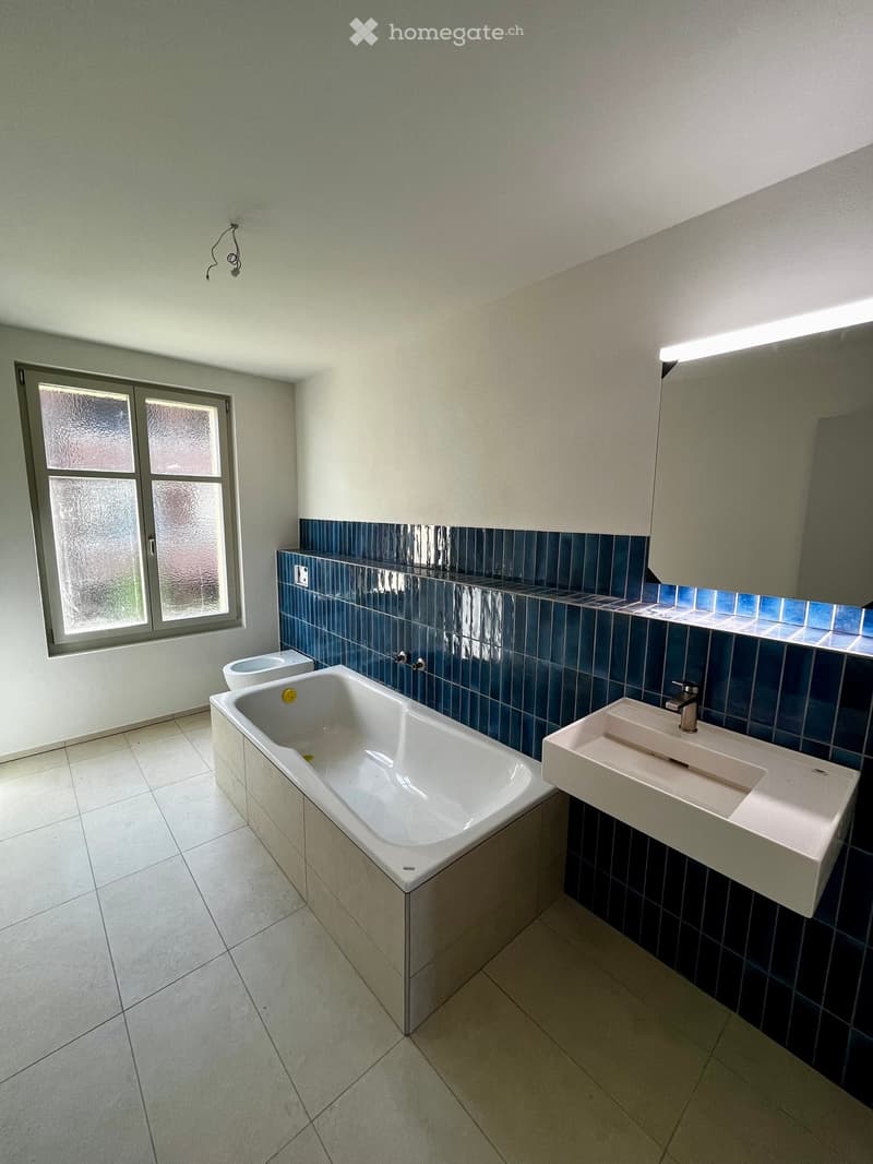 Modern bathroom with a bathtub, sink, and toilet. Blue tiles on the walls above the fixtures, white tiles on the floor, and a large window providing natural light.
