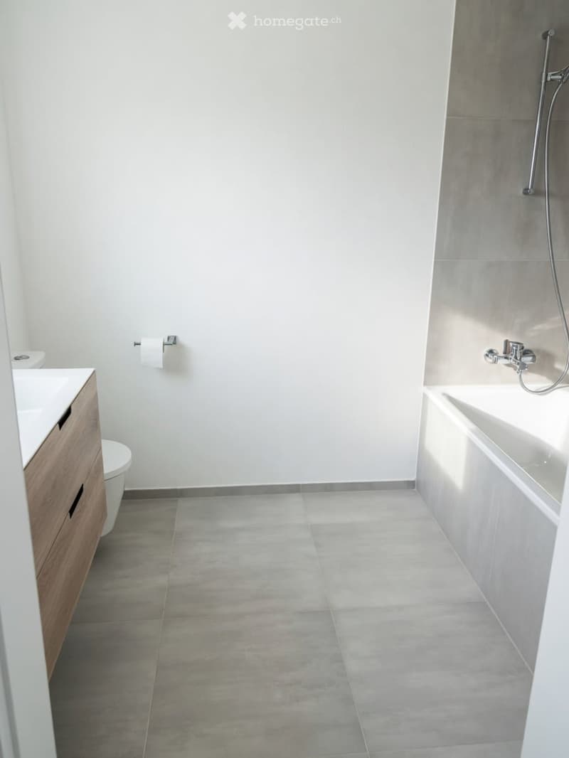 Modern bathroom with a white tiled wall, grey tiled floor, a wooden vanity with a sink, a toilet, and a bathtub with a chrome faucet.