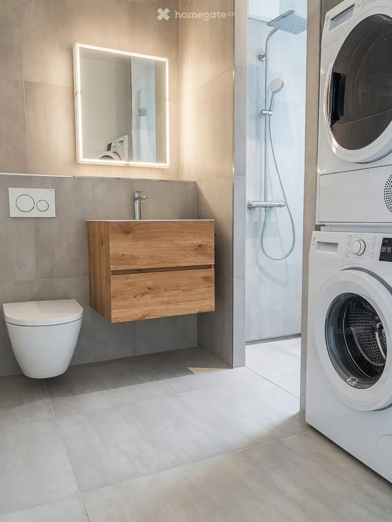 Modern bathroom with a wooden sink cabinet, toilet, mirror with LED lighting, and a shower area with a washing machine and dryer unit.