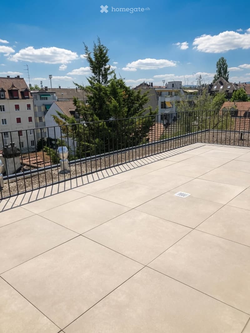 Roof terrace with concrete tiles, metal railing, and a view of surrounding buildings and trees.