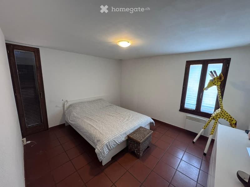 Bedroom with a bed, a small stool, a giraffe toy, and a table near the window. The room has tiled flooring and white walls.